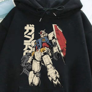 May include: Black hooded sweatshirt featuring a colorful illustration of a robot with a shield and Japanese text. The robot is primarily white, blue, red, and yellow. The sweatshirt has a front pocket and a drawstring hood.