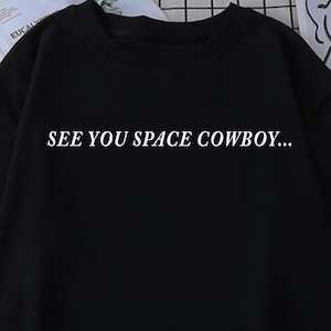 Op de afbeelding: Zwart T-shirt met korte mouwen en de witte tekst "SEE YOU SPACE COWBOY...". Het shirt is gemaakt van een zacht materiaal en heeft een ronde hals. Het shirt is een casual kledingstuk.