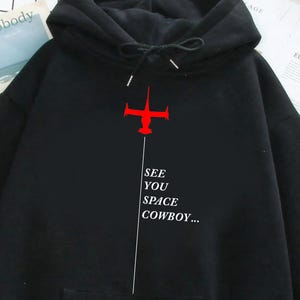 Pode incluir: Camisola preta com capuz, bolso frontal e cordão. O design apresenta um gráfico vermelho em forma de cruz com uma linha branca e o texto "SEE YOU SPACE COWBOY..." em branco. A camisola é feita de um material macio.