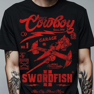 Op de afbeelding: Zwart t-shirt met een rood grafisch ontwerp. Het ontwerp bevat de tekst "Space Cowboy Garage" en "Swordfish II", samen met een ruimteschip illustratie. Extra tekst omvat "Since 1998" en "Alba City, Planet Mars".