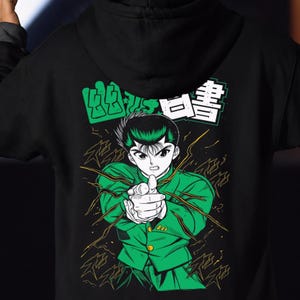 Peut inclure: Un sweat à capuche noir avec une impression graphique au dos. Le motif représente un personnage d'anime en veste verte pointant vers l'avant, entouré d'effets d'éclairs jaunes et de texte japonais dans des blocs verts et blancs au-dessus.