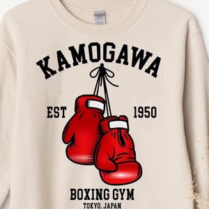 May include: A beige crewneck sweatshirt featuring a graphic of two red boxing gloves hanging by their laces. The text reads KAMOGAWA, EST 1950, BOXING GYM, and TOKYO. JAPAN in black lettering. The design is centered on the front of the garment.