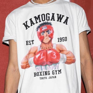 Puede incluir: Una camiseta blanca de manga corta con el gráfico de un boxeador con casco rojo y guantes de boxeo rojos. El texto dice KAMOGAWA EST 1950 BOXING GYM TOKYO, JAPAN. El boxeador tiene el pelo oscuro y lleva una camiseta sin mangas blanca.