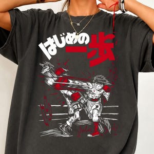 Puede incluir: Una camiseta gris carbón con un estampado gráfico de un boxeador en movimiento. El diseño incluye texto en japonés blanco y rojo, una ilustración en blanco y negro de un luchador lanzando un puñetazo y detalles rojos en los guantes.
