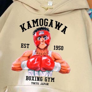 May include: A beige hooded sweatshirt featuring a graphic print of a boxer wearing red headgear and red boxing gloves. The text reads KAMOGAWA, EST 1950, BOXING GYM, and TOKYO, JAPAN. The boxer has the numbers 1 and 3 on the gloves.