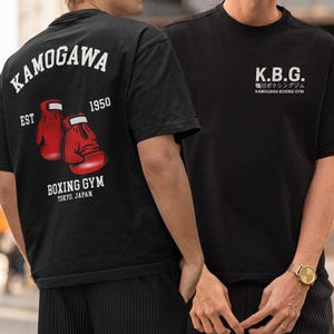 Puede incluir: Una camiseta negra con un gráfico en la espalda de dos guantes de boxeo rojos, el texto KAMOGAWA EST 1950 BOXING GYM TOKYO. JAPAN, y un gráfico en el pecho con K.B.G. y el texto japonés 鴨川ボクシングジム KAMOGAWA BOXING GYM.