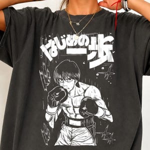 Puede incluir: Una camiseta gris carbón con un estampado gráfico blanco de un boxeador estilo manga en posición de combate con guantes y pantalones cortos. Sobre el personaje, el texto japonés "はじめの一歩" está impreso en una fuente blanca.