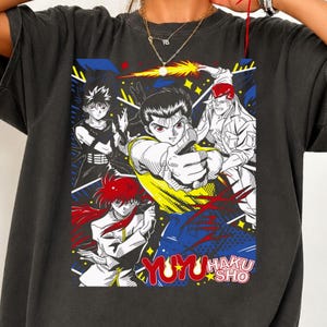 Pode incluir: Uma t-shirt de manga curta cinzento-carvão com uma impressão gráfica de quatro personagens de anime de YuYu Hakusho. O design inclui linhas marcadas, detalhes em vermelho, amarelo e azul, e o texto YU YU HAKUSHO em vermelho e branco.