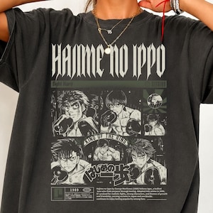 Puede incluir: Una camiseta gris carbón con un gran estampado gráfico con el texto "HAJIME NO IPPO". El diseño incluye varias ilustraciones en blanco y negro estilo manga de un boxeador en varias poses, junto con texto en japonés y el año 1989.