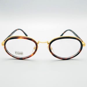 Könnte beinhalten: Ovale Brille mit goldfarbenem Rahmen und Schildpattmuster auf den Rändern. Die Bügel sind dunkelbraun und grün. Der Markenname "Gianfranco Ferre Lunettes" ist auf dem Glas sichtbar.