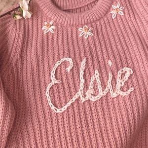 May include: A pink knitted jumper with white embroidered flowers and the name "Ellie" on the front.
