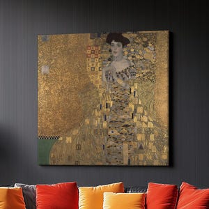 May include: A large framed artwork featuring a portrait of a woman in a gold and patterned dress. The artwork hangs above a black leather sofa with orange and red throw pillows. A gray ottoman and a round coffee table are in front of the sofa.
