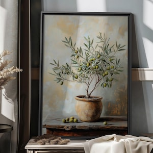 May include: A framed painting depicting an olive tree in a terracotta pot, placed on a wooden table. The artwork showcases green olives and leaves, against a neutral backdrop. The scene is lit by natural light, creating a calm atmosphere.