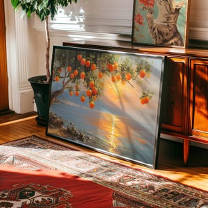 May include: A framed landscape painting depicts an orange tree laden with fruit, overlooking a body of water at sunset. The artwork is in a black frame and leans against a wooden cabinet. A patterned rug and a potted plant are in the foreground.