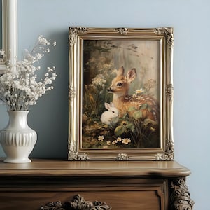 May include: A framed painting of a fawn and a white rabbit amidst wildflowers and foliage. The artwork is presented in a gold, ornate frame. The painting is displayed on a wooden surface, adding a classic touch.