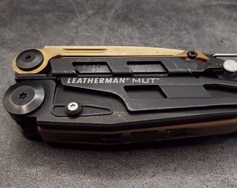 Può includere: Un multiutensile Leatherman MUT nero con un manico dorato. Lo strumento ha una varietà di accessori, tra cui un coltello, una sega e un cacciavite. Lo strumento è etichettato "LEATHERMAN MUT".