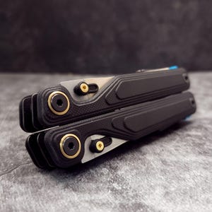 Pode incluir: Uma multi-ferramenta preta com uma pega texturizada e detalhes dourados. A ferramenta tem uma lâmina prateada e um detalhe azul na extremidade. A ferramenta está fechada e pousada numa superfície cinzenta.