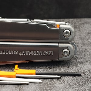 Może przedstawiać: Srebrne wielofunkcyjne narzędzie z napisem "LEATHERMAN. SURGE" na boku. Narzędzie ma czarną rękojeść i pomarańczowe akcenty.