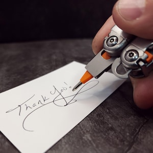 Puede incluir: Una multiherramienta plateada y naranja escribe "Thank You!" en una tarjeta blanca. La herramienta tiene una punta de destornillador extendida, y el texto está en cursiva negra. El fondo es una superficie oscura y texturizada.