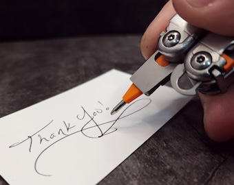 Può includere: Un multiutensile argentato e arancione sta scrivendo "Thank You!" su una carta bianca. L'utensile ha una punta di cacciavite estesa e il testo è in corsivo nero. Lo sfondo è una superficie scura e strutturata.