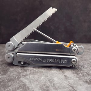 Op de afbeelding: Een zilveren multitool met een gedeeltelijk geopend zaagblad. De tool heeft een oranje accent en de merknaam "LEATHERMAN WAVE" is zichtbaar. De tool is ontworpen voor diverse taken.