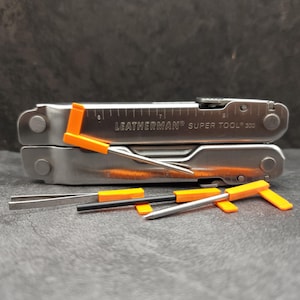 May include: A silver Leatherman Super Tool 300 multi-tool with an integrated ruler marked in inches. The tool is accompanied by several small, detachable accessories featuring bright orange plastic components, including a pick and tweezers.