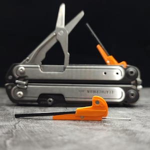 May include: A silver Leatherman Arc multi-tool with deployed metal scissors and a black and orange accessory piece. A separate, smaller black and orange tool component with a thin metal pin is positioned in the foreground.