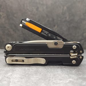 以下が含まれることがあります： 黒いLeatherman ARCマルチツール。アームが一部展開されており、T8およびT6と刻印されたオレンジ色のハンドル付きビットドライバーが見える。本体は黒で、銀色の金具、ポケットクリップ、側面にUSA MagnaCutの刻印がある。