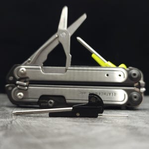 May include: A silver Leatherman ARC multi-tool with deployed scissors and a yellow-handled driver bit, positioned behind a small black metal component with a thin silver pin extending from its side, all featuring a metallic finish.