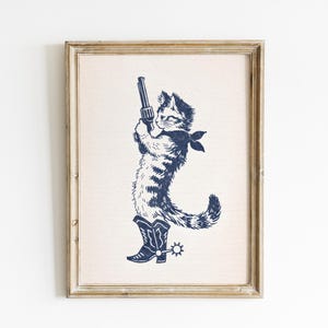 Peut inclure: Impression encadrée avec une illustration bleu marine d'un chat en tenue de cow-boy, tenant un pistolet. Le chat porte un bandana et des bottes de cowboy avec des éperons. L'œuvre est sur fond beige clair.