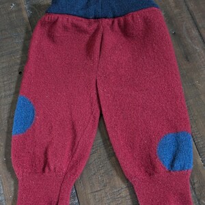 May include: A pair of knit pants in a deep red color featuring a dark blue elasticized waistband and matching dark blue circular patches on each knee area. The pants have ribbed cuffs at the bottom of each leg and a soft, textured knit construction.