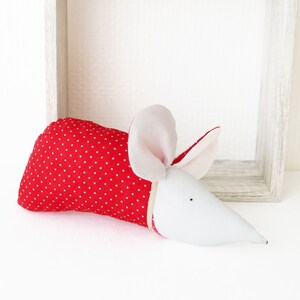 May include: A red and white polka dot fabric mouse with a white felt face and ears. The mouse is lying on a white surface.