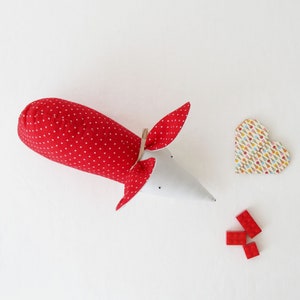 May include: A red and white polka dot fabric mouse with a white felt body and a red felt nose. The mouse is lying on a white surface with a heart-shaped paper craft and red plastic building blocks.