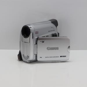 Puede incluir: Una videocámara digital plateada Canon MV901 con formato Mini DV. El dispositivo cuenta con un objetivo de zoom óptico de 25x, zoom digital de 800x y una pantalla abatible que muestra el logotipo de Canon, el sistema PAL y el modelo MV901.