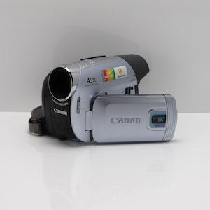 May include: A silver and black Canon camcorder featuring a 45x advanced zoom lens, a flip-out LCD screen, and MiniDV MD235 branding. The device includes a Canon Video Lens, a 2.7 inch widescreen indicator, and a Quick Start button on the body.