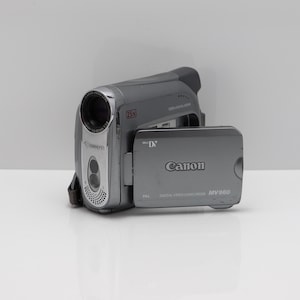 May include: A grey Canon MV960 digital video camcorder with a flip-out LCD screen. The device features a 25x optical zoom lens, Mini DV branding, and PAL system designation. The Canon logo is visible on the side panel and the front lens housing.