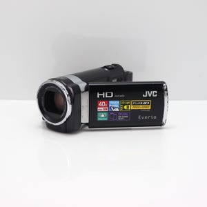 Op de afbeelding: Een zwarte JVC Everio camcorder met een uitklapbaar scherm met iconen voor HD, 40x optische zoom, touchpanel, LED-licht, Full HD 1920x1080, dubbele SD-kaartsleuf en stabilisator. Het apparaat heeft een lens met een zilverkleurige rand.