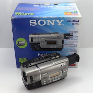 Può includere: Una videocamera Sony Handycam TR427E color argento e nero con obiettivo zoom digitale 80x, funzionalità NightShot e controlli di messa a fuoco manuale, posizionata davanti alla scatola di imballaggio originale blu e bianca con testo.