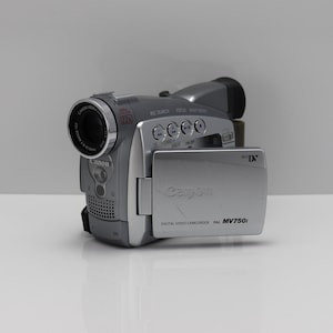 Op de afbeelding: Een zilverkleurige Canon MV750i digitale videocamera met een 22x optische zoomlens, een uitklapbaar LCD-scherm met Mini DV-branding en diverse bedieningsknoppen op de bovenkant van de behuizing. Het apparaat heeft een luidsprekerrooster.