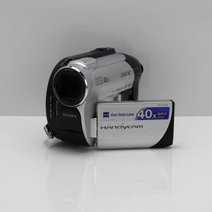 May include: A silver and black Sony Handycam camcorder featuring a Carl Zeiss lens with 40x optical zoom. The device has a flip-out screen displaying the text Carl Zeiss Lens 40x Optical Zoom and Handycam. The camera body includes Sony branding.