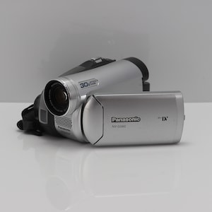 Op de afbeelding: Een zilverkleurige Panasonic NV-GS60 MiniDV-camcorder met een 30x optische zoomlens. Het apparaat heeft een uitklapbaar scherm met het Panasonic-logo en de MiniDV-merknaam aan de zijkant. De behuizing is compact met zwarte accenten.