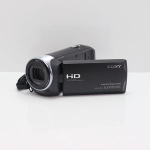 May include: A black Sony Handycam video camera with a flip-out screen. The device features a Zeiss lens, HD AVCHD branding, and 9.2 Mega Pixels text on the side. The camera body is compact with a silver lens ring and a black wrist strap attached.