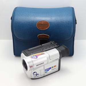 May include: A silver Samsung camcorder featuring a 22x optical zoom lens, 880x digital zoom, and Easy-Q button, positioned in front of a blue textured carrying case with a brown leather-like oval patch displaying the Samsung brand name.