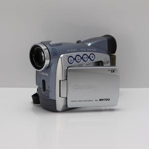 Puede incluir: Una videocámara digital Canon MV700 de color plata y gris con ranura para cinta MiniDV. El dispositivo cuenta con un objetivo de zoom óptico de 18x, una pantalla LCD abatible y botones de control REC SEARCH, FOCUS y NIGHT MODE en la parte superior.