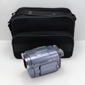 May include: A blue Sony Handycam video camera with a VideoHi8 label, featuring a side-hinged screen and various control buttons, positioned in front of a black rectangular carrying bag with a top handle and zippered compartments.