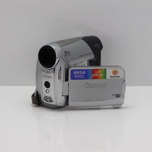Pode incluir: Uma câmara de vídeo MiniDV Canon prateada e preta com lente de zoom ótico de 35x e ecrã LCD panorâmico rebatível de 2,7 polegadas. O ecrã apresenta o texto Mega Pixel, 2.7 inch Wide LCD, Quick Start e o logótipo Canon em letras prateadas.