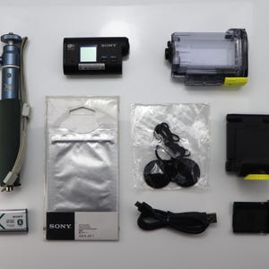 May include: A Sony action camera kit including a blue monopod, a black camera body, a clear waterproof housing with a yellow latch, a battery, a USB cable, a black mount, a plastic bag with two circular adhesive mounts, and a Sony AKA-AF1 package.