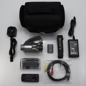 May include: A Sony camcorder kit including a black carrying bag, a silver and black camcorder, a battery, a power adapter with a cord, a remote control, a black strap, a clear plastic housing, and a multi-colored AV cable in a plastic bag.