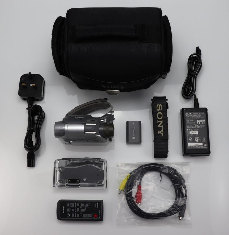 May include: A Sony camcorder kit including a black carrying bag, a silver and black camcorder, a battery, a power adapter with a cord, a remote control, a black strap, a clear plastic housing, and a multi-colored AV cable in a plastic bag.