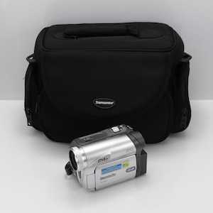 Op de afbeelding: Een zilverkleurige Panasonic digitale camcorder met een 24x optische zoomlens en een zwarte Samsonite draagtas. De camcorder heeft een klein scherm met blauwe en witte tekstlabels, een zilveren knop aan de bovenkant en een zwarte batterijhouder.